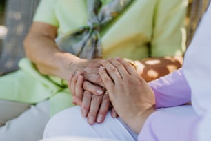 A close-up of caregiver consoling senior woman and touching her hand when sitting on bench in park in summer.