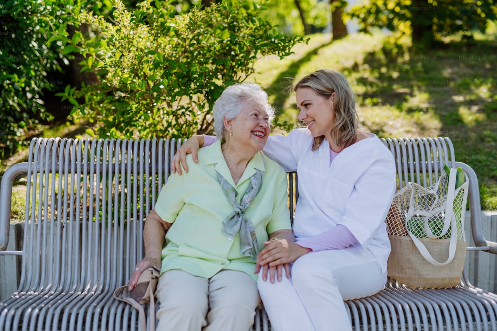 A caregiver with senior woman sitting on bench in park in summer, looking at camera.