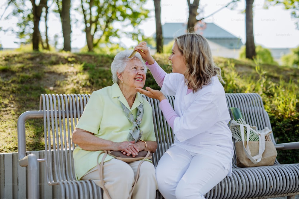 A caregiver helping senior woman to comb hair and make hairstyle when sitting on bench in park in summer.