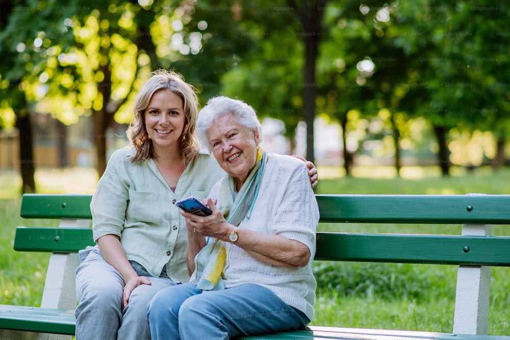 An adult granddaguhter helping her grandmother to use cellphone when sitting on bench in park in summer.