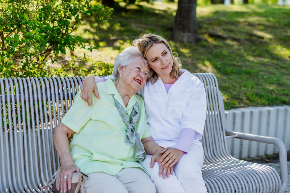 A caregiver with senior woman sitting on bench in park in summer, embracing.