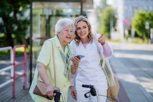 Una badante con una donna anziana che cammina nel parco con la borsa della spesa, aiutando con l'uso di uno smartphone.