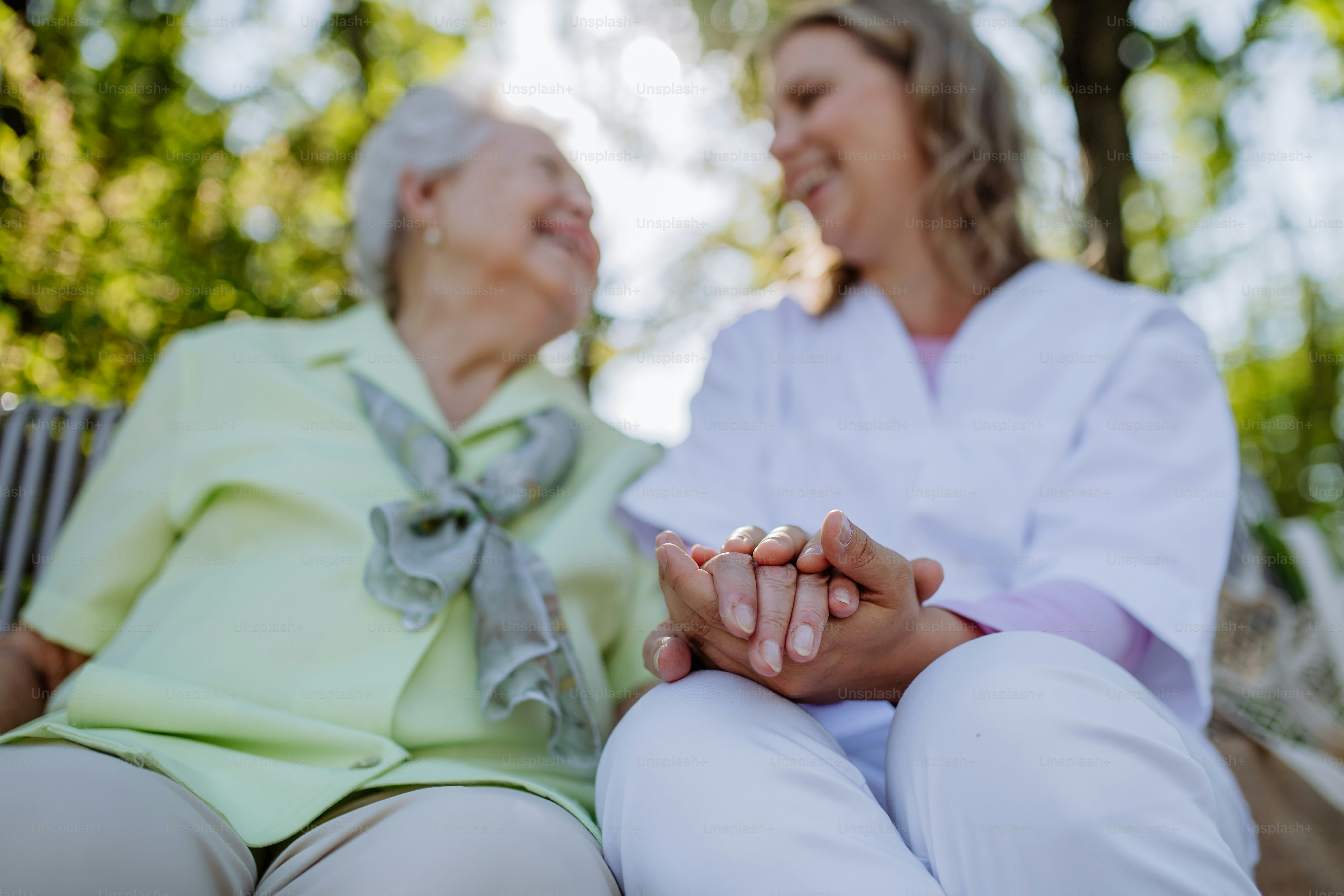 A low angle view of caregiver consoling senior woman and touching her hand when sitting on bench in park in summer.