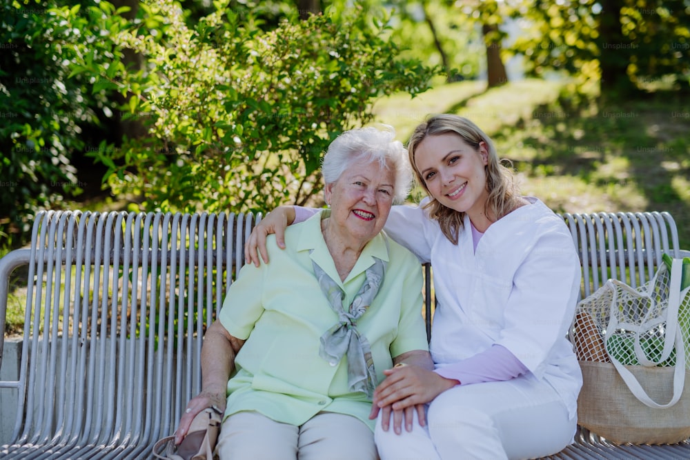 A caregiver with senior woman sitting on bench in park in summer, looking at camera.