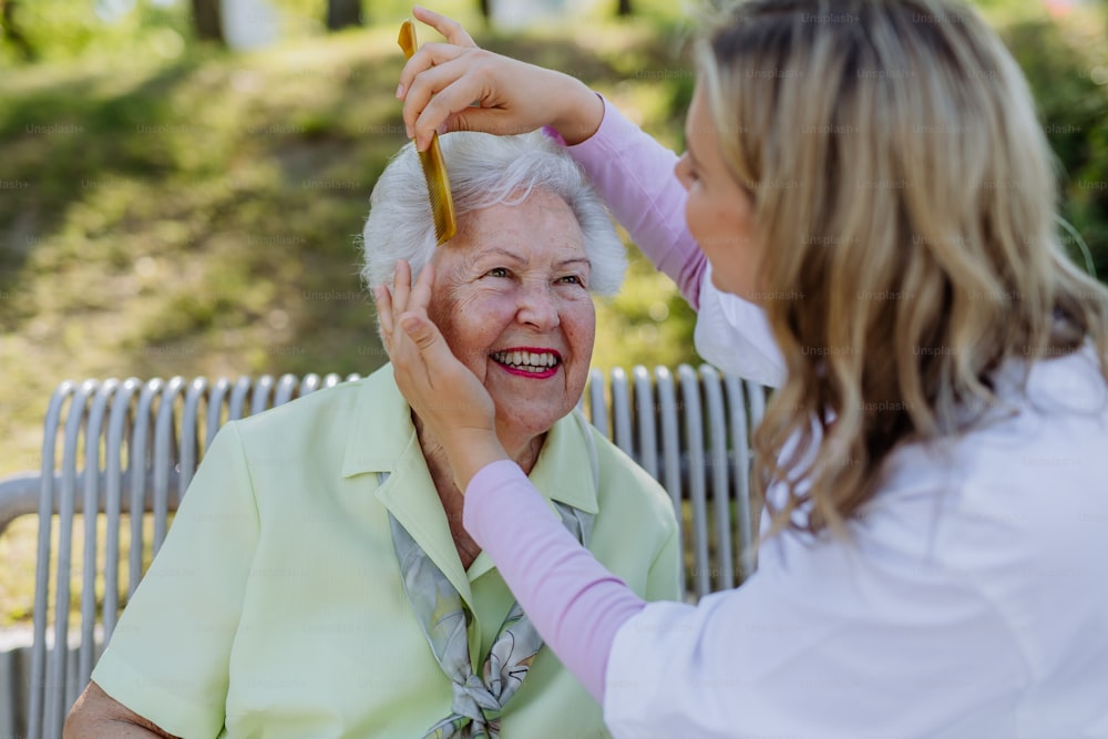 A caregiver helping senior woman to comb hair and make hairstyle when sitting on bench in park in summer.