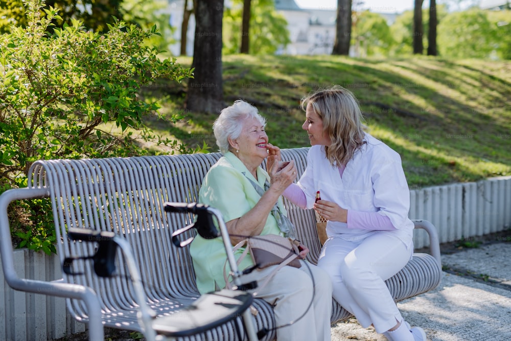 A caregiver helping senior woman to apply lipstick when sitting on bench in park in summer.