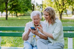 An adult granddaguhter helping her grandmother to use cellphone when sitting on bench in park in summer.