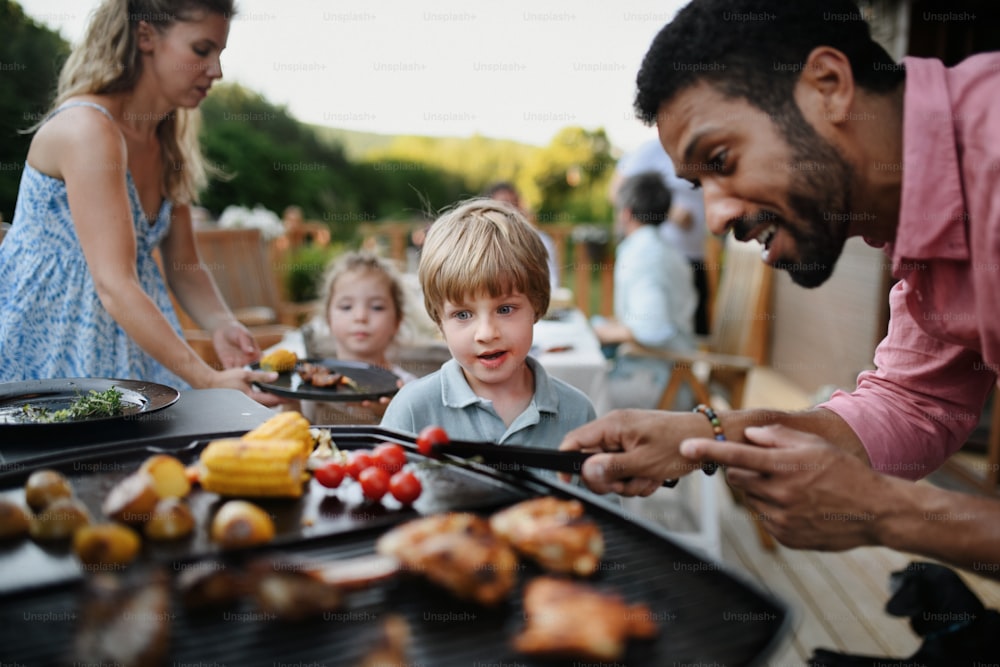 A father grilling meat and vegetable on grill during family summer garden party.