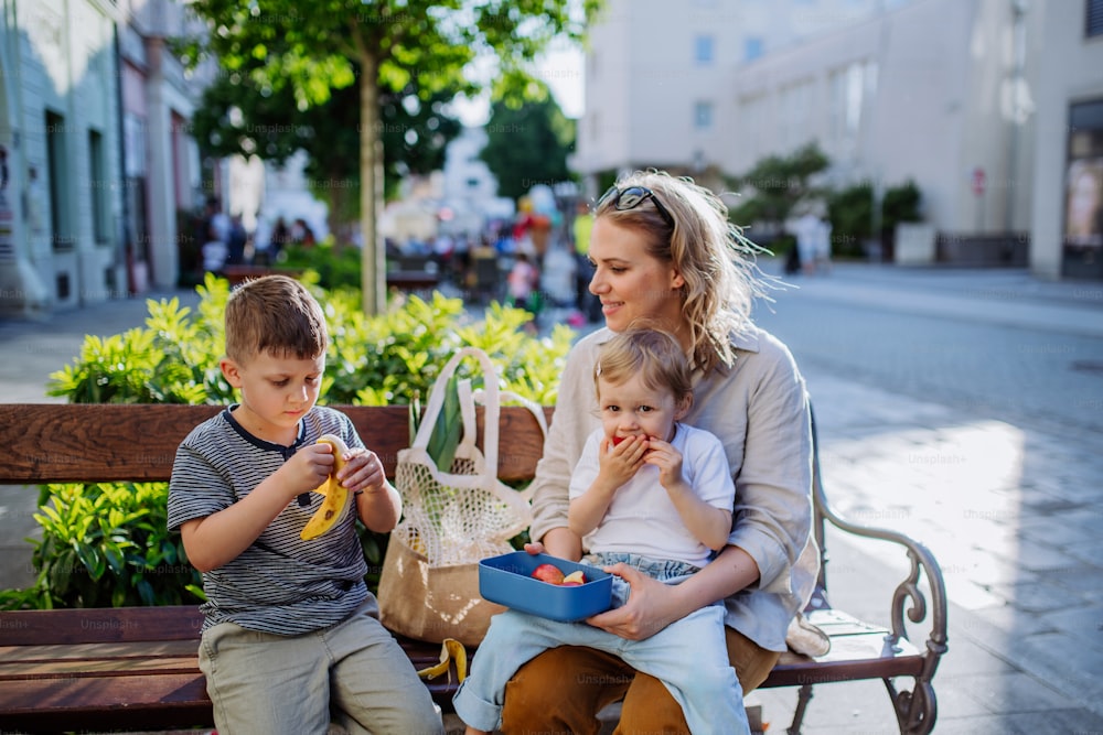 A young mother with little kids resting on bench in city and eating fruit snack.