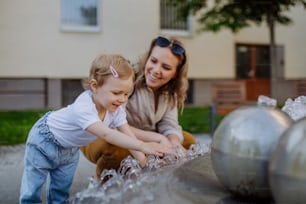 A young mother playing with her kid by fountain in city in summer.