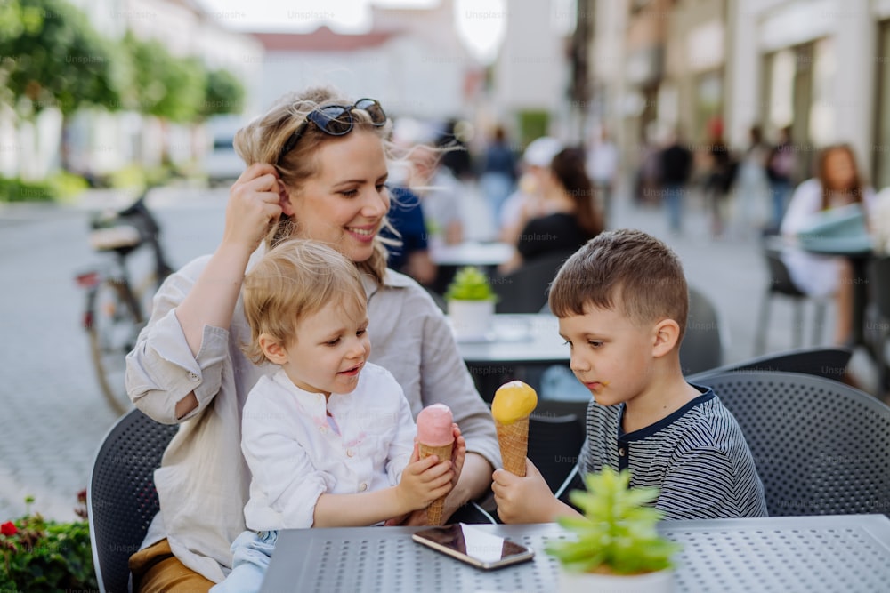 A young mother taking selfie with her kids eating ice-cream in cafe outdoors in street in summer.