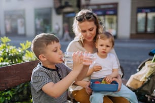 Una giovane madre con bambini piccoli seduti sulla panchina in città in estate, mangiando snack di frutta e acqua potabile.