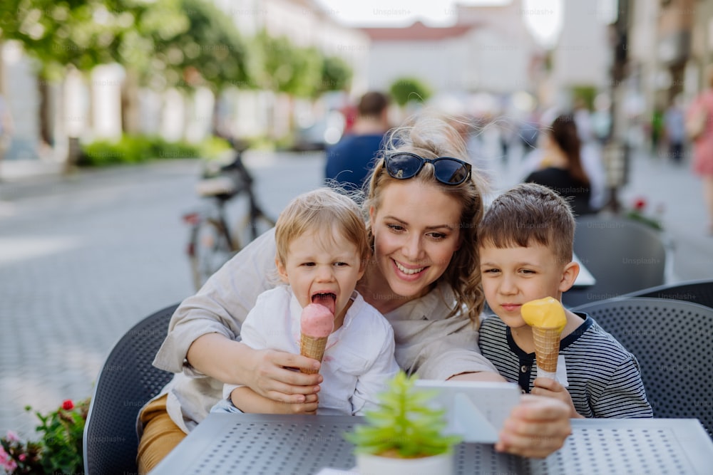 Eine junge Mutter, die im Sommer ein Selfie mit ihren Kindern beim Eisessen im Café im Freien auf der Straße macht.