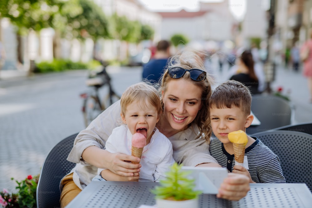 A young mother taking selfie with her kids eating ice-cream in cafe outdoors in street in summer.