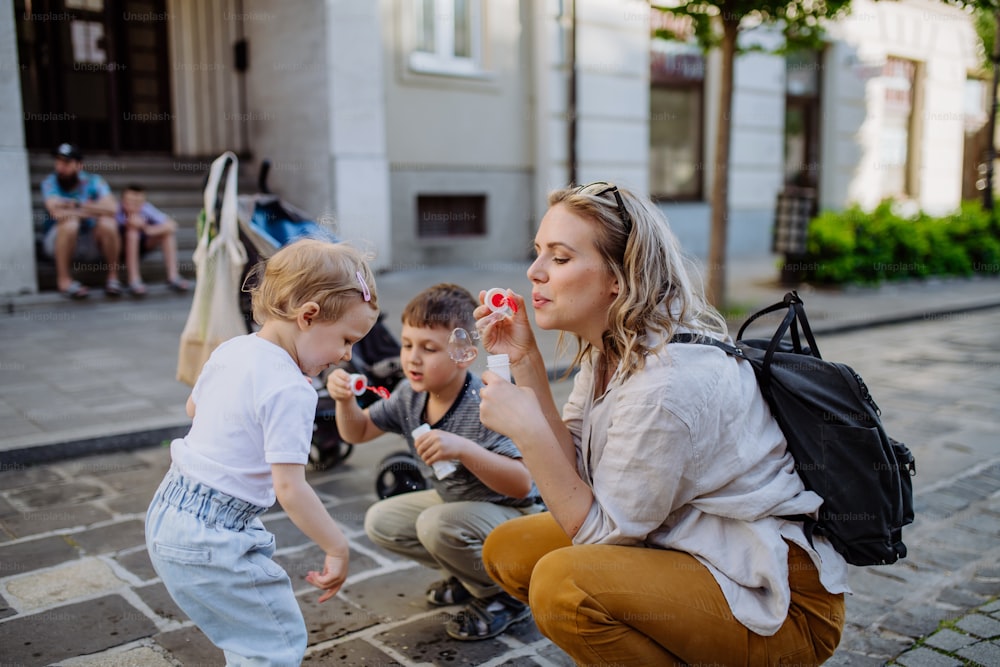 A young mother playing with her kids, blowing bubbles in city street in summer.