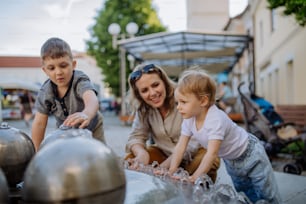 Una giovane madre che gioca con i suoi bambini con la fontana in strada della città in estate.