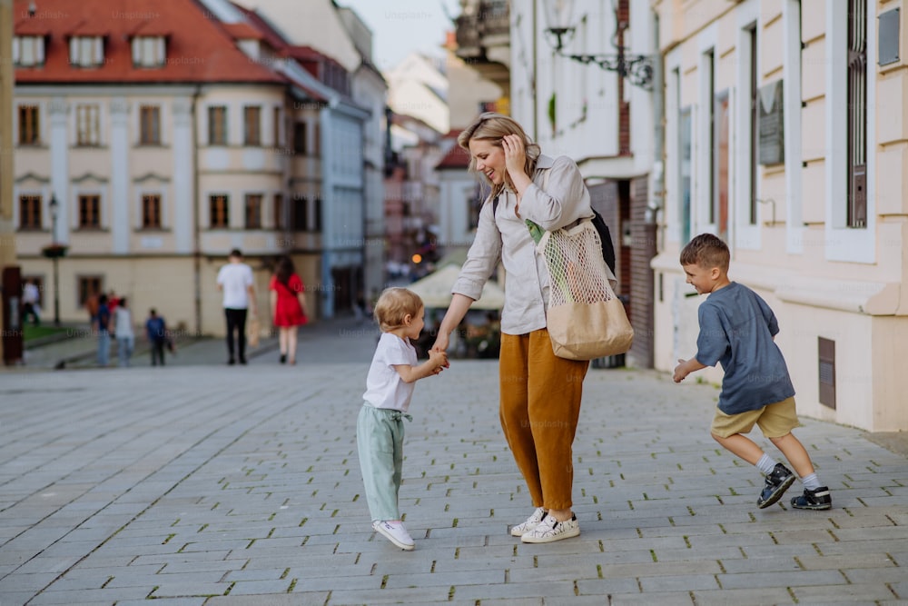 Una joven madre con bolsa de compras cero residuos tomada de la mano con sus hijos y caminando por la calle de la ciudad