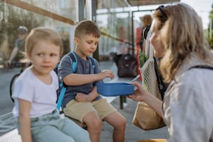 小さな子供を持つ若い母親が街のバス停で待っていて、果物のおやつを食べています。