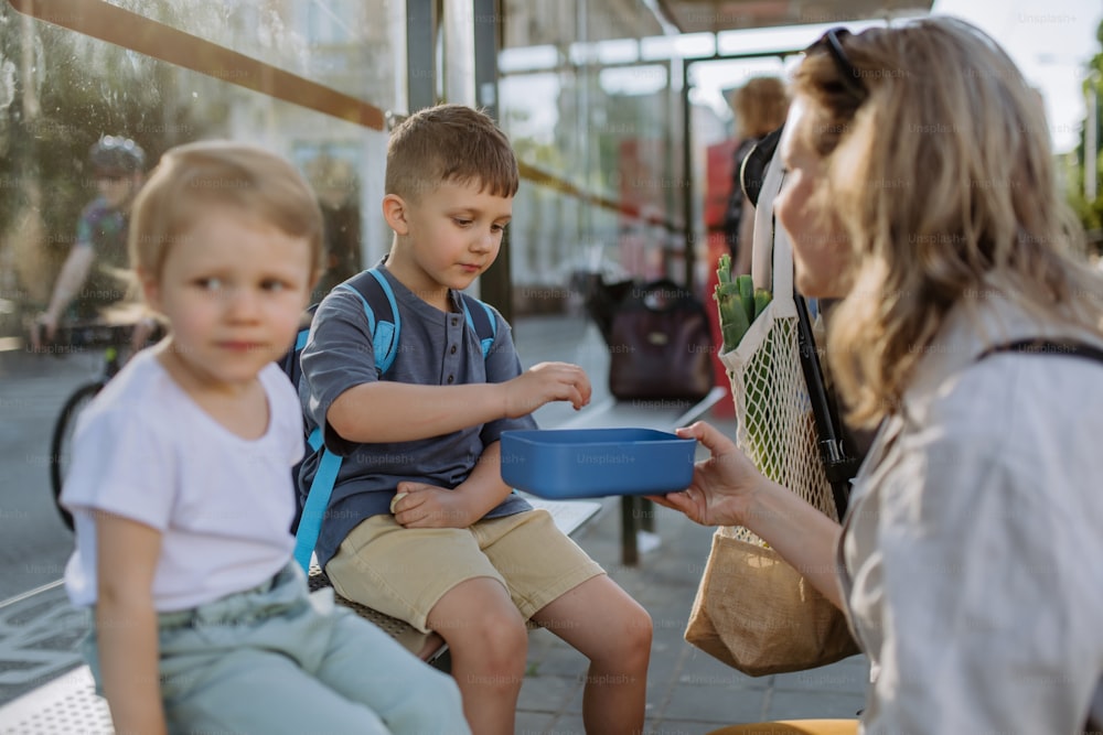 Una joven madre con niños pequeños esperando en una parada de autobús en la ciudad y comiendo bocadillos de frutas.