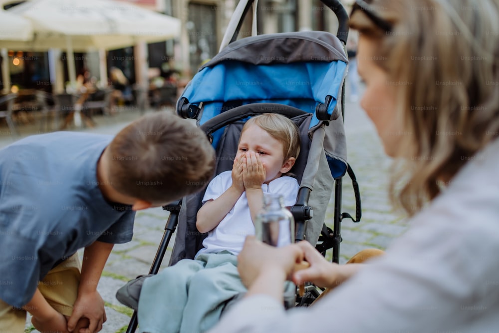 A mother and little brother calming a crying girl sitting in a stroller while walking down the street