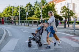 Eine junge Mutter mit Zero-Waste-Einkaufstasche, die Händchen mit ihrem Kind und Kinderwagen hält, wenn sie auf der Straße der Stadt spazieren geht