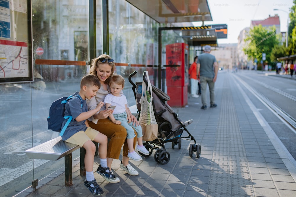 Una joven madre con niños pequeños esperando en una parada de autobús en la ciudad, desplazándose en el teléfono móvil.