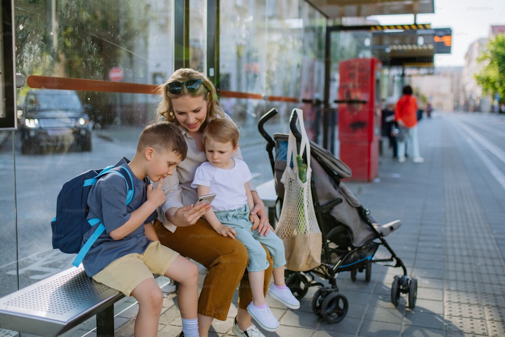 A young mother with little kids waiting on bus stop in city, scrolling on mobile phone.