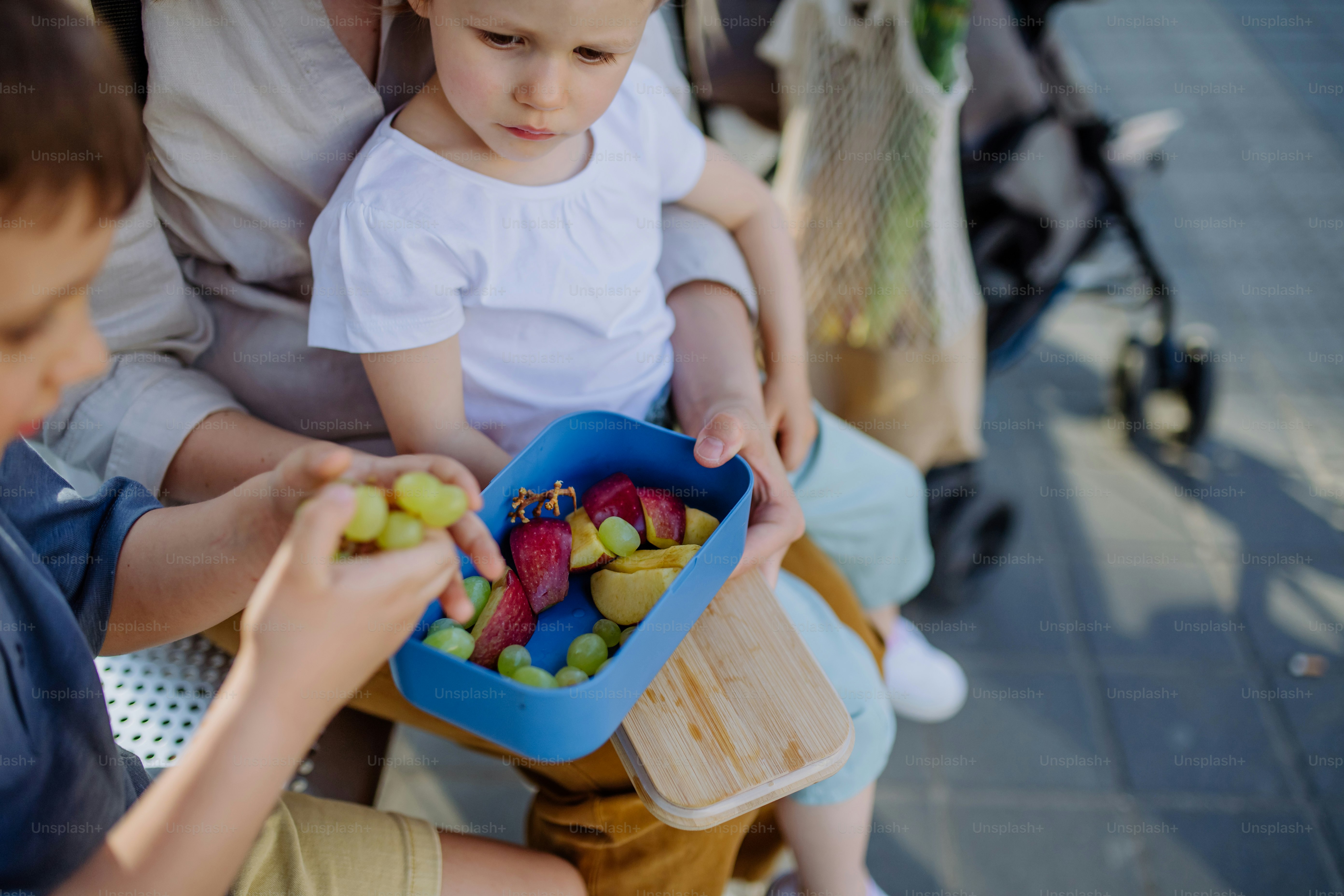 Uma jovem mãe com crianças pequenas esperando no ponto de ônibus na cidade e comendo lanche de frutas
