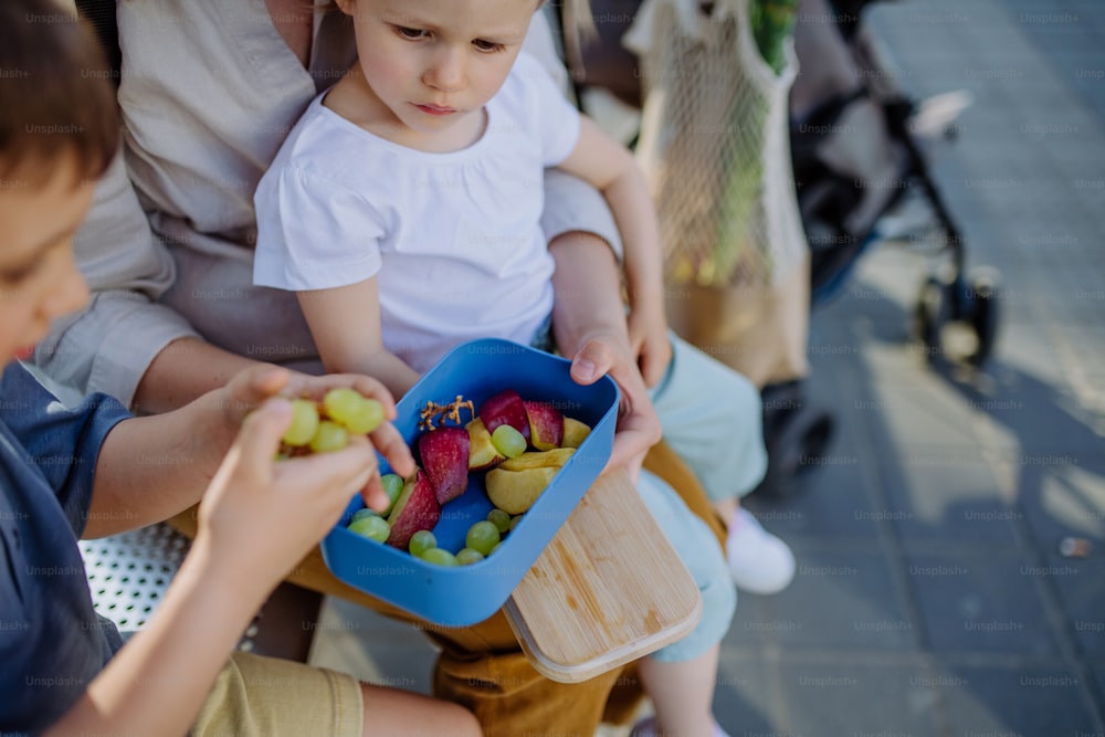 Una joven madre con niños pequeños esperando en una parada de autobús en la ciudad y comiendo bocadillos de frutas.