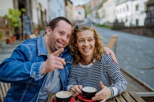 Un portrait d’un jeune homme heureux atteint du syndrome de Down avec sa mère assise dans un café en plein air et en train de parler.
