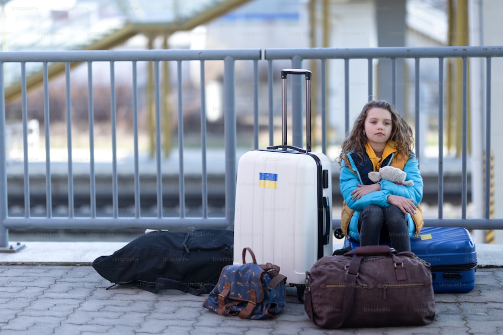 A sad Ukrainian immigrant child with luggage waiting at train station, Ukrainian war concept.