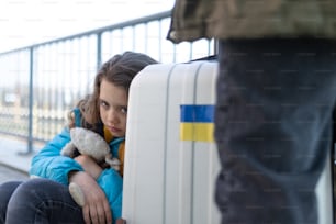 A sad Ukrainian immigrant child with luggage waiting at train station, Ukrainian war concept.
