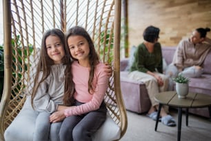 Happy little sisters sitting in wicker rattan hang chair indoors in conservatory, with mother and grandmother at background.
