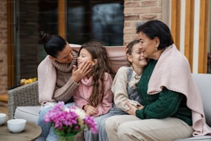 Two happy sisters with a mother and grandmother sitting wrapped in blanket outdoors in patio in autumn
