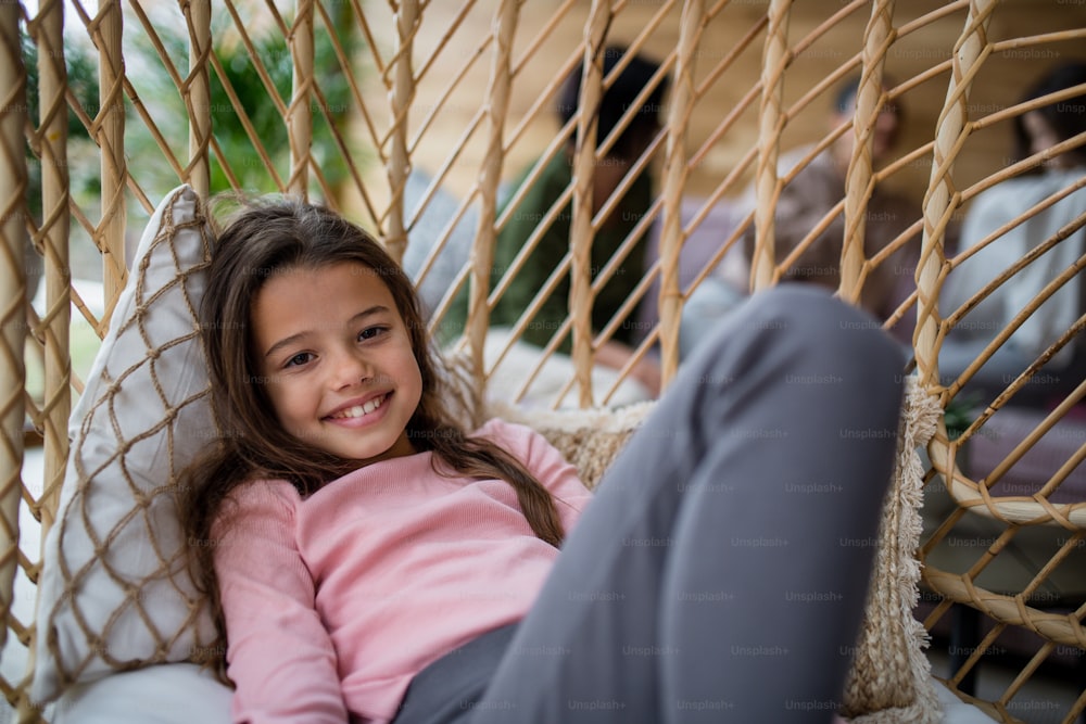 A happy little girl sitting in wicker rattan hang chair outdoors in patio in autumn, looking at camera