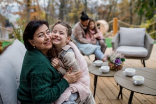 A happy little girl with grandmother sitting wrapped in blanket outdoors in patio in autumn