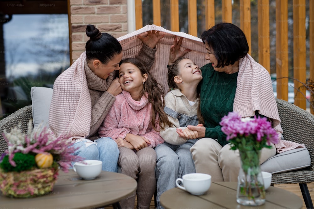 Two happy sisters with a mother and grandmother sitting wrapped in blanket outdoors in patio in autumn
