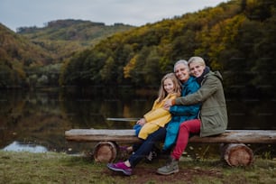 A small girl with mother and grandmother sitting on bench and looking at camera outoors by lake.