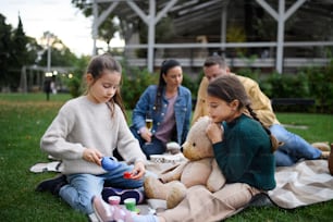 Happy little sisters sitting on blanket and playing with teddy bear on picnic with parents outdoor in park