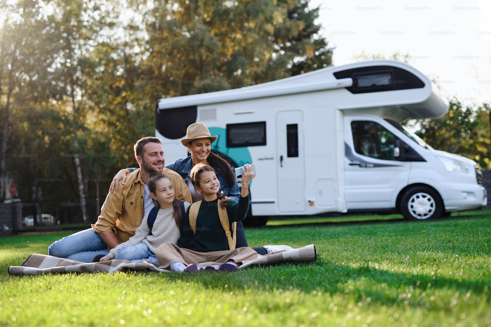 Une jeune famille heureuse avec deux enfants prenant un selfie avec caravane en arrière-plan à l’extérieur.