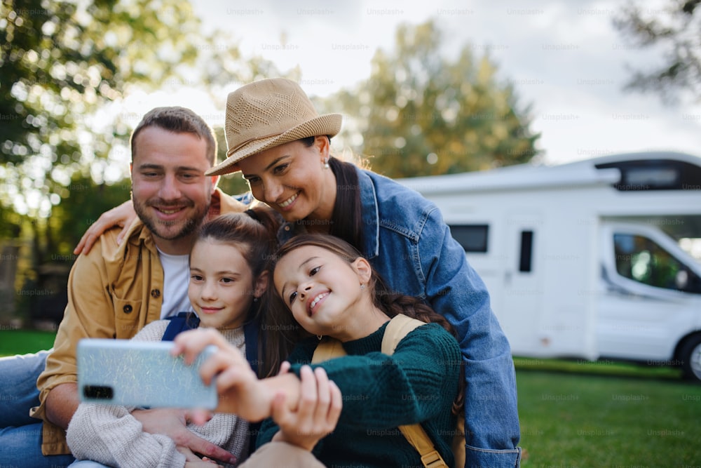 Une jeune famille heureuse avec deux enfants prenant un selfie avec caravane en arrière-plan à l’extérieur.