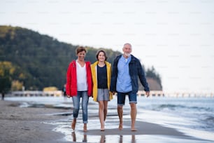 A senior couple holding hands with their preteen granddaughter and walking on sandy beach.