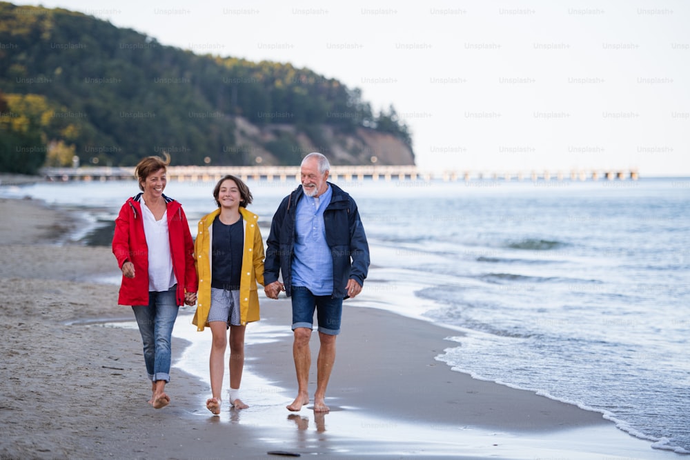 A senior couple holding hands with their preteen granddaughter and walking on sandy beach.