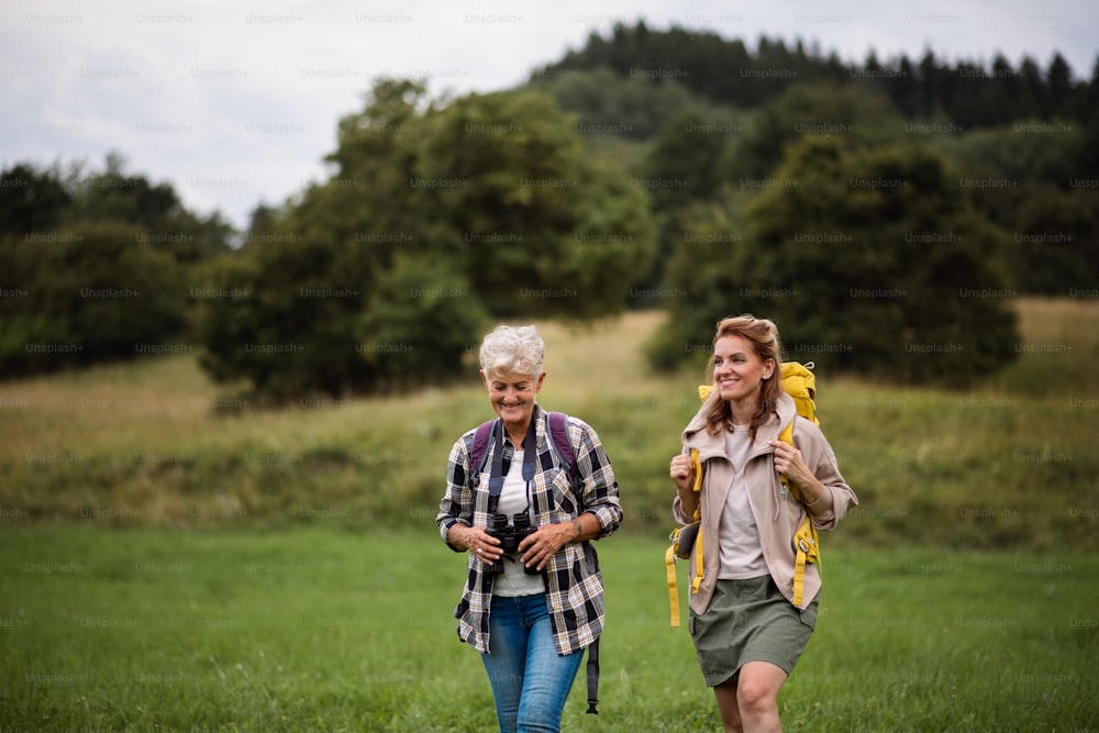 Una mujer mayor activa con binoculares caminando con su hija adulta al aire libre en la naturaleza. Una mujer adulta mediana feliz con bastones de trekking caminando con una madre mayor activa al aire libre en la naturaleza.