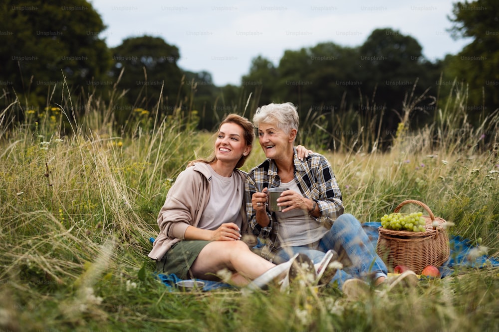 Eine glückliche ältere Mutter und eine erwachsene Tochter, die draußen in der Natur sitzen und picknicken und reden.