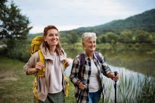 A happy senior woman with trekking poles hiking with adult daughter outdoors in nature.