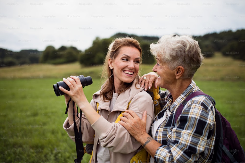 A happy senior mother hiker embracing with adult daughter holding binoculars outdoors in nature