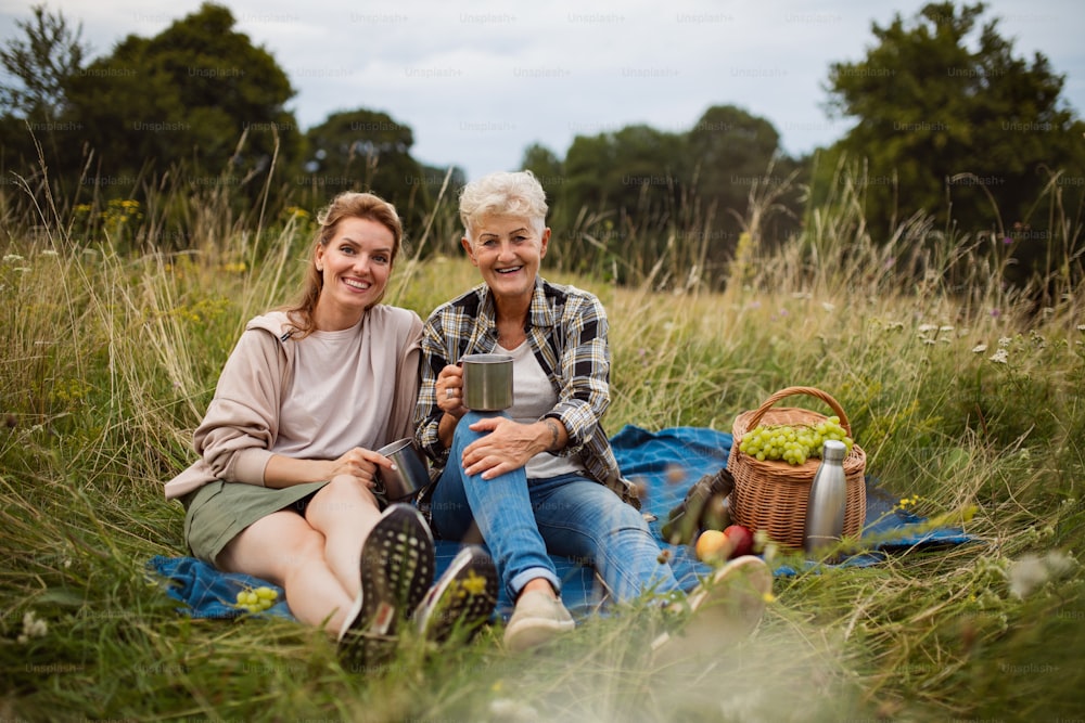 A happy senior mother and adult daughter sitting and having picnic outdoors in nature, looking at camera.