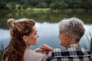 A happy senior mother with adult daughter sitting in front of lake outdoors in nature.