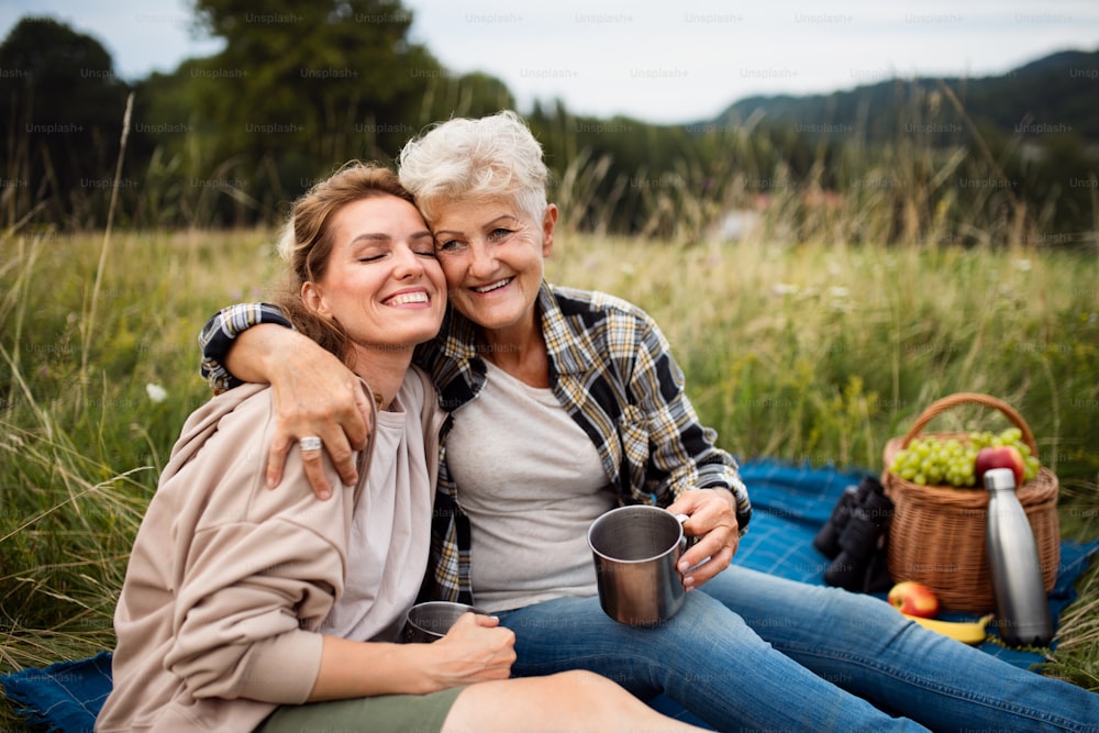 A happy senior mother embracing adult daughter when sitting and having picnic outdoors in nature.
